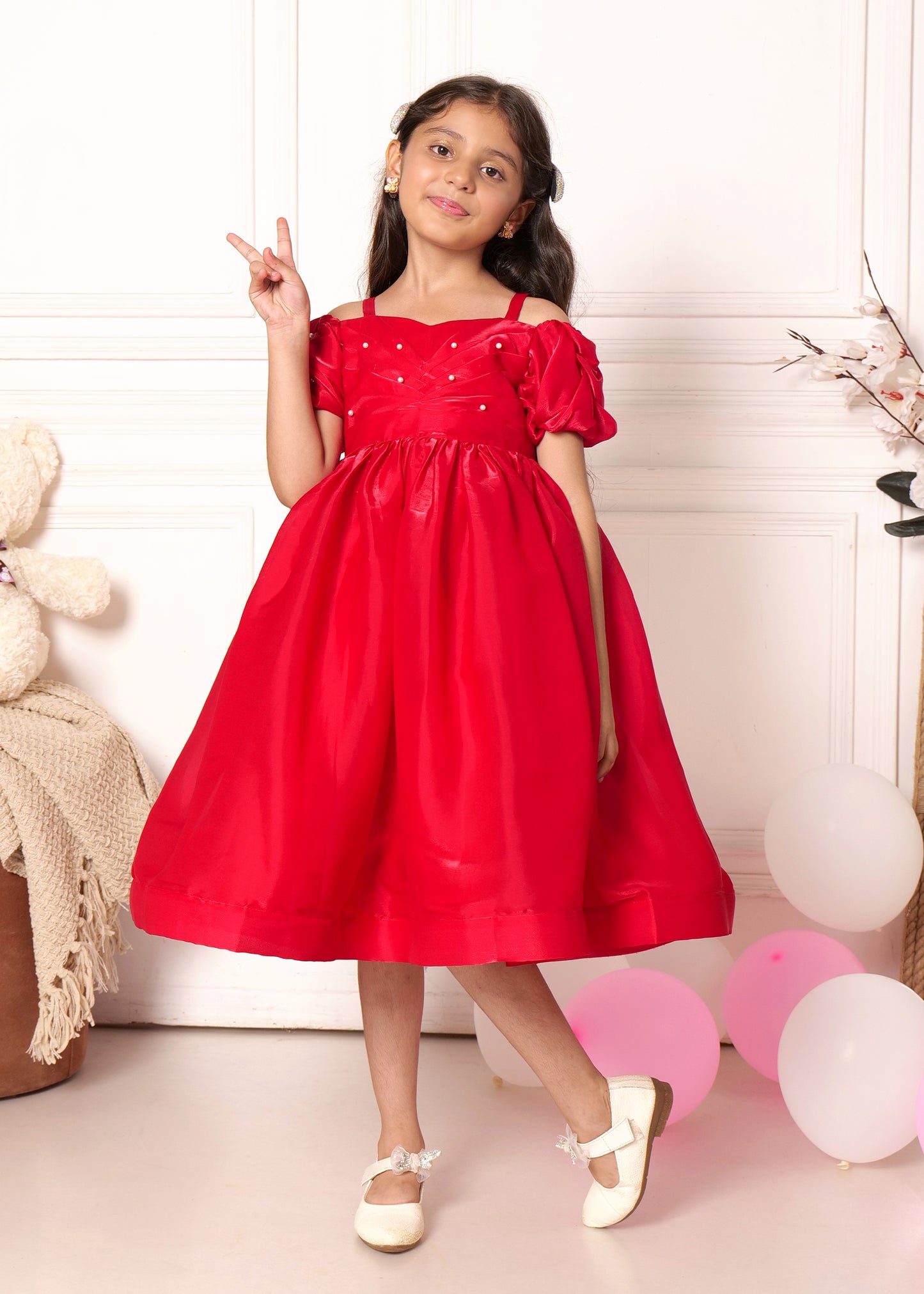 "Smiling girl in a beautiful red party dress with puff sleeves and pearl accents, standing next to balloons, making a peace sign in a joyous celebration."