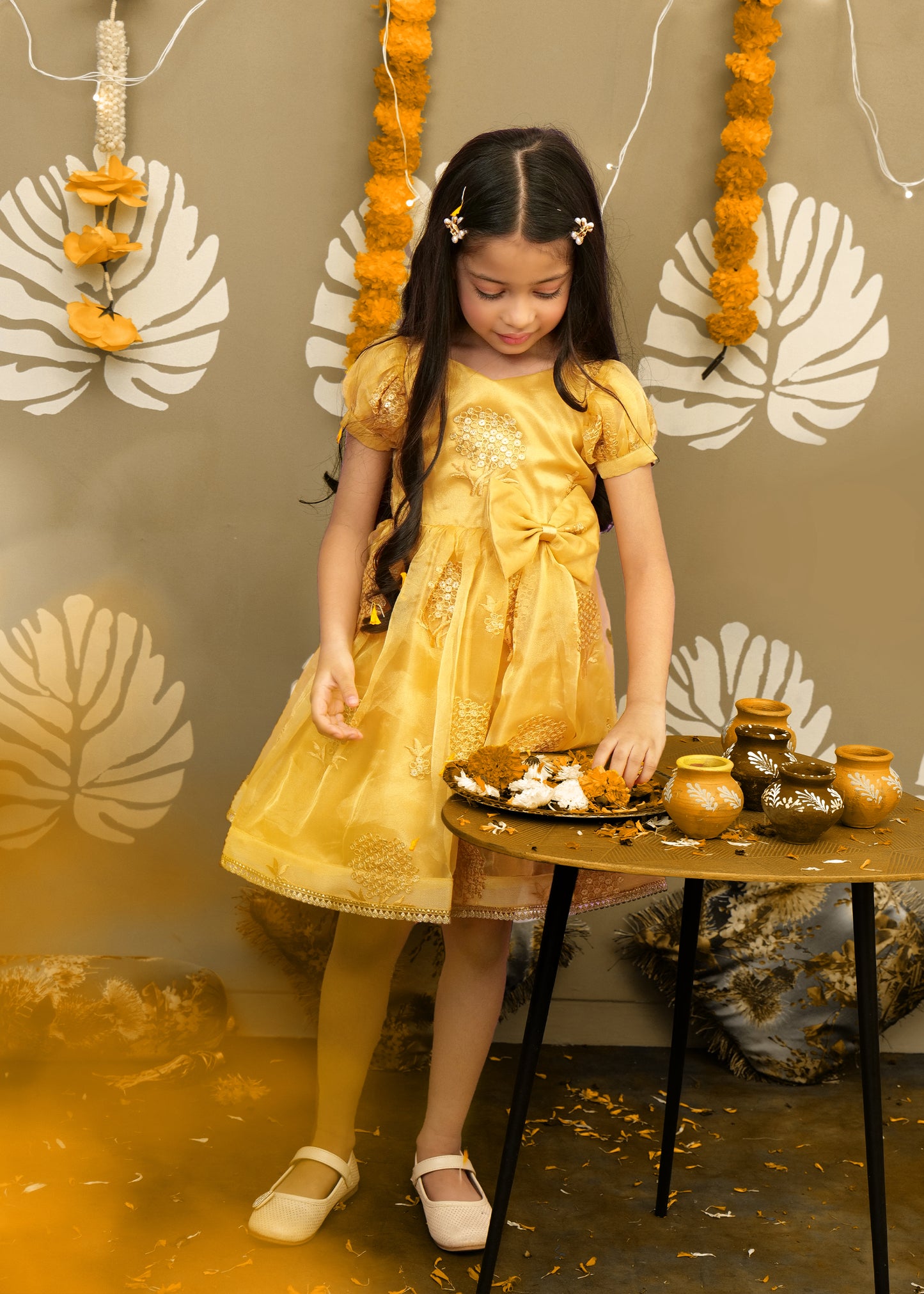 Girl in a golden party dress organizing marigold flowers and decorative clay pots during a traditional Indian festival.