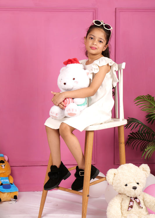 Little girl in a white ruffled dress seated on a chair, holding a stuffed teddy bear, wearing black boots, surrounded by playful decor in a vibrant pink setting