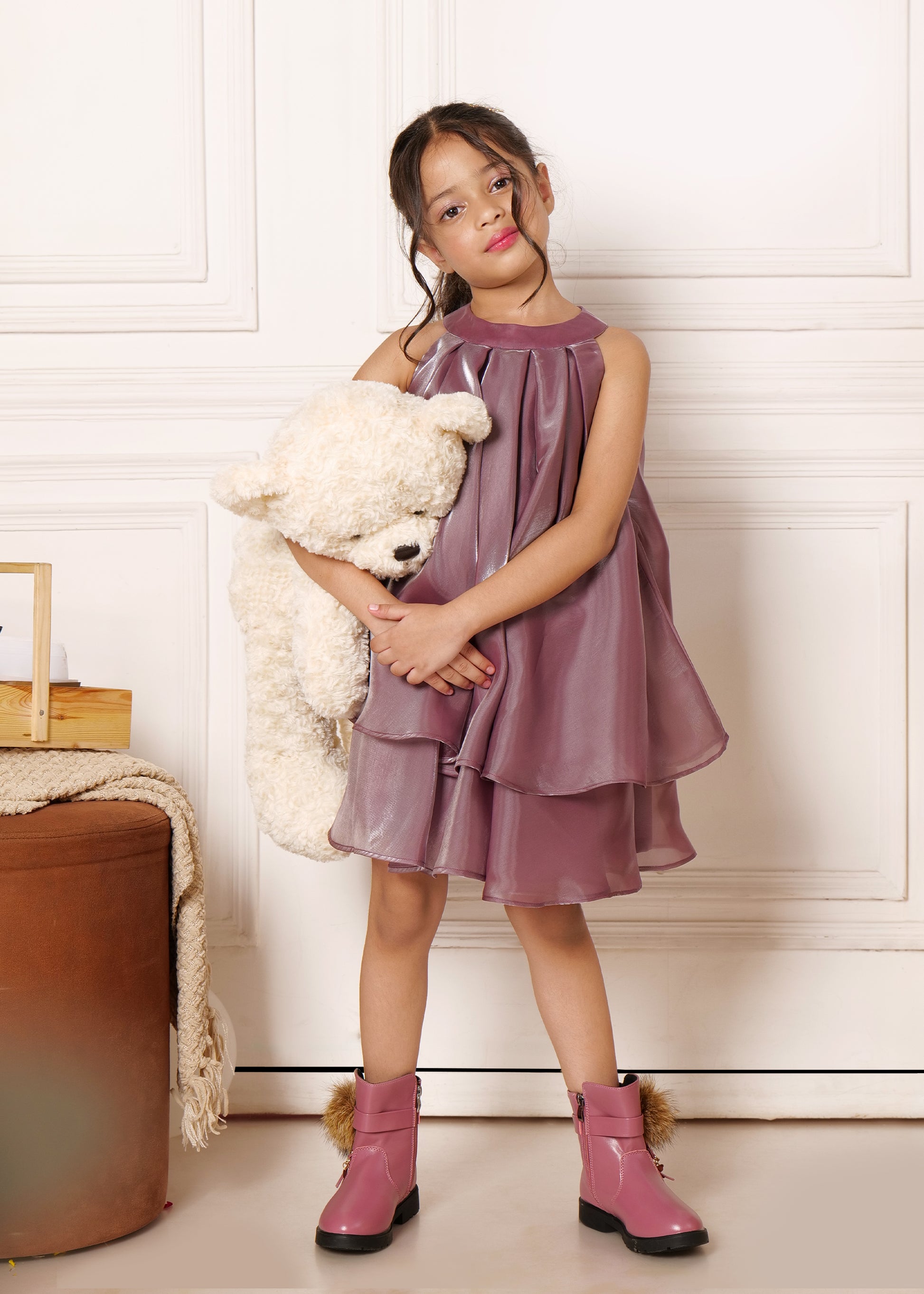 Adorable child holding a teddy bear, dressed in a shiny purple satin dress and pink boots, styled for a cute and trendy look