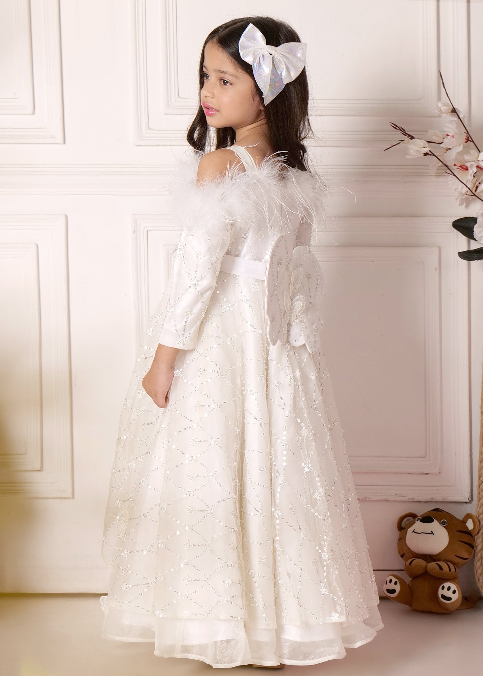 Side view of a young girl wearing a white embellished gown with butterfly accents and a large bow, styled with a soft white hair bow.