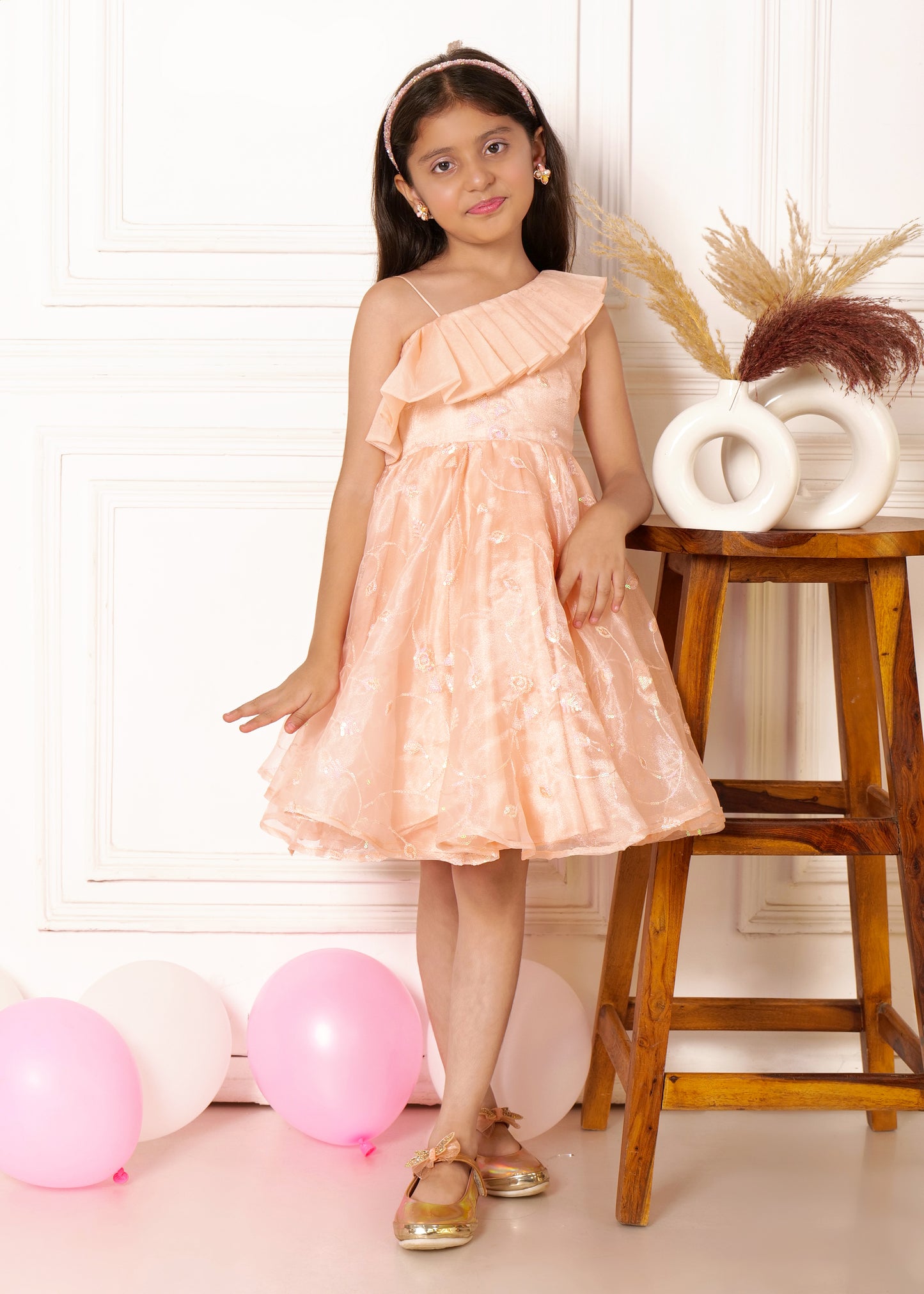 "A young girl wearing a peach-colored dress with a one-shoulder ruffle design and floral details, standing by a wooden stool with balloons in the background, smiling gently at the camera."