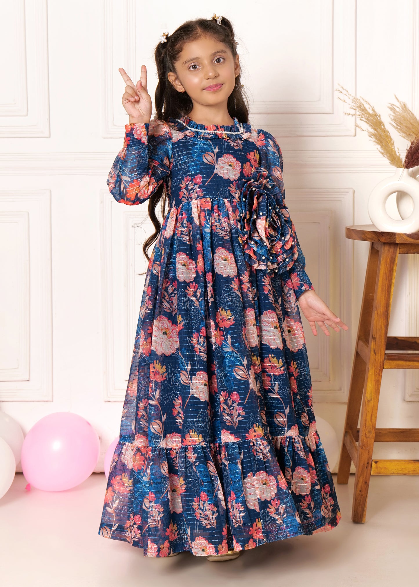 Girl dressed in a floral long-sleeved maxi dress, striking a peace sign pose, surrounded by pastel balloons, perfect for children's spring and summer fashion campaigns.