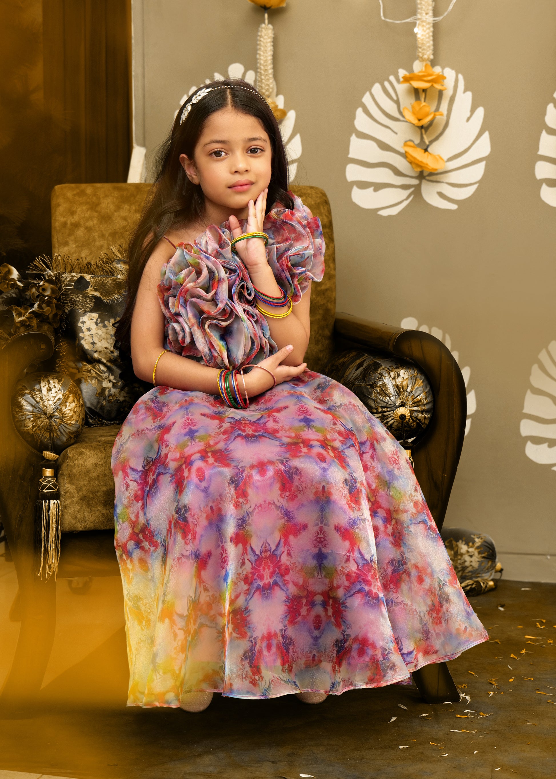  Girl sitting elegantly in a chair wearing a colorful lehenga and ruffle blouse, adorned with traditional bangles.