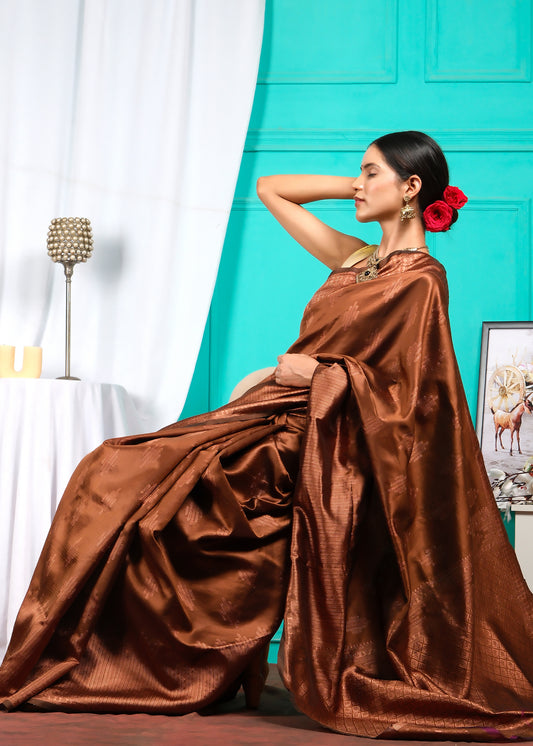 Model posing gracefully in a brown silk saree, styled with statement ethnic jewelry, seated in an elegant indoor setup.