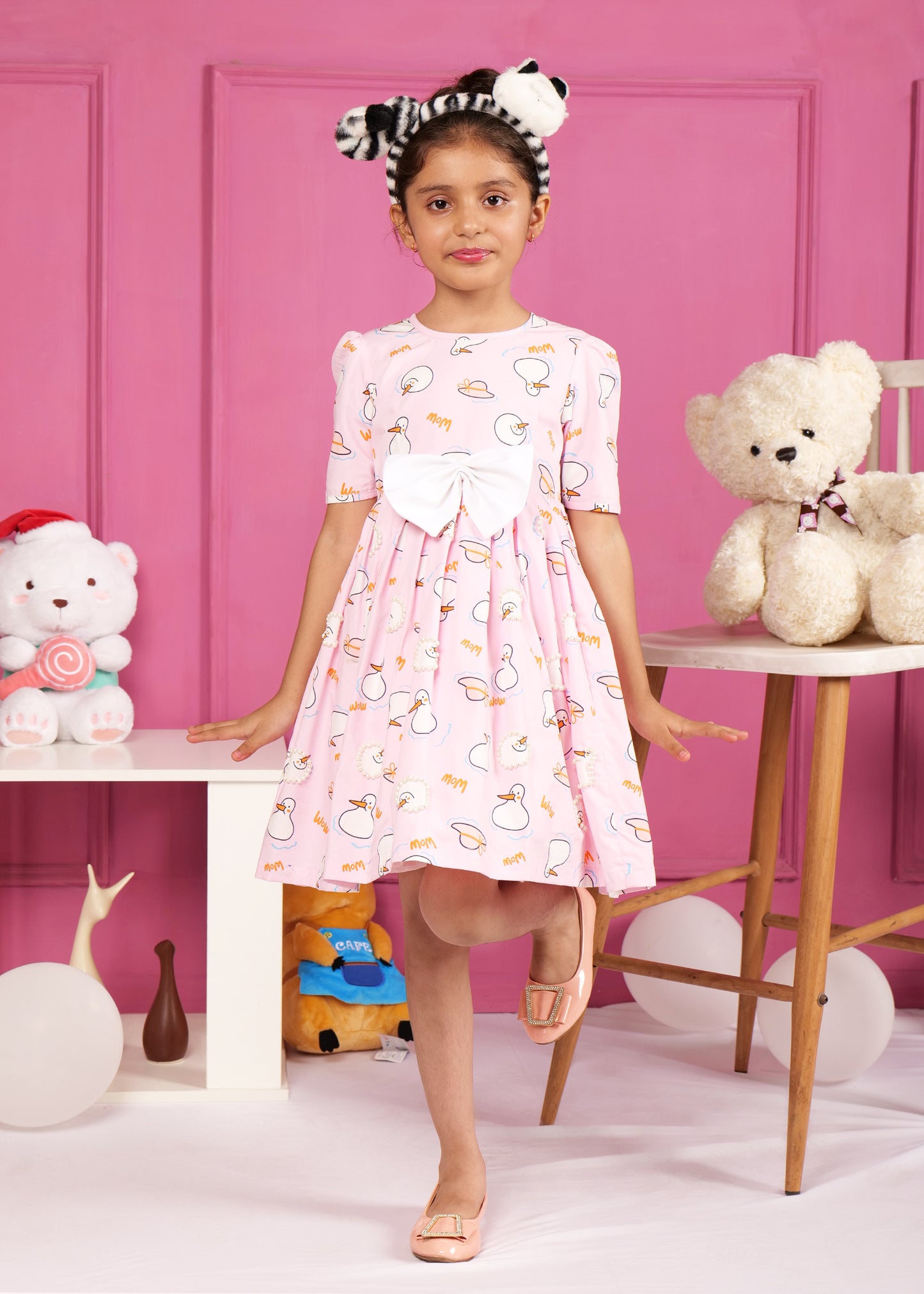 Girl in a charming pink dress with cute swan prints, striking a casual pose in front of a playful backdrop with soft toys and balloons