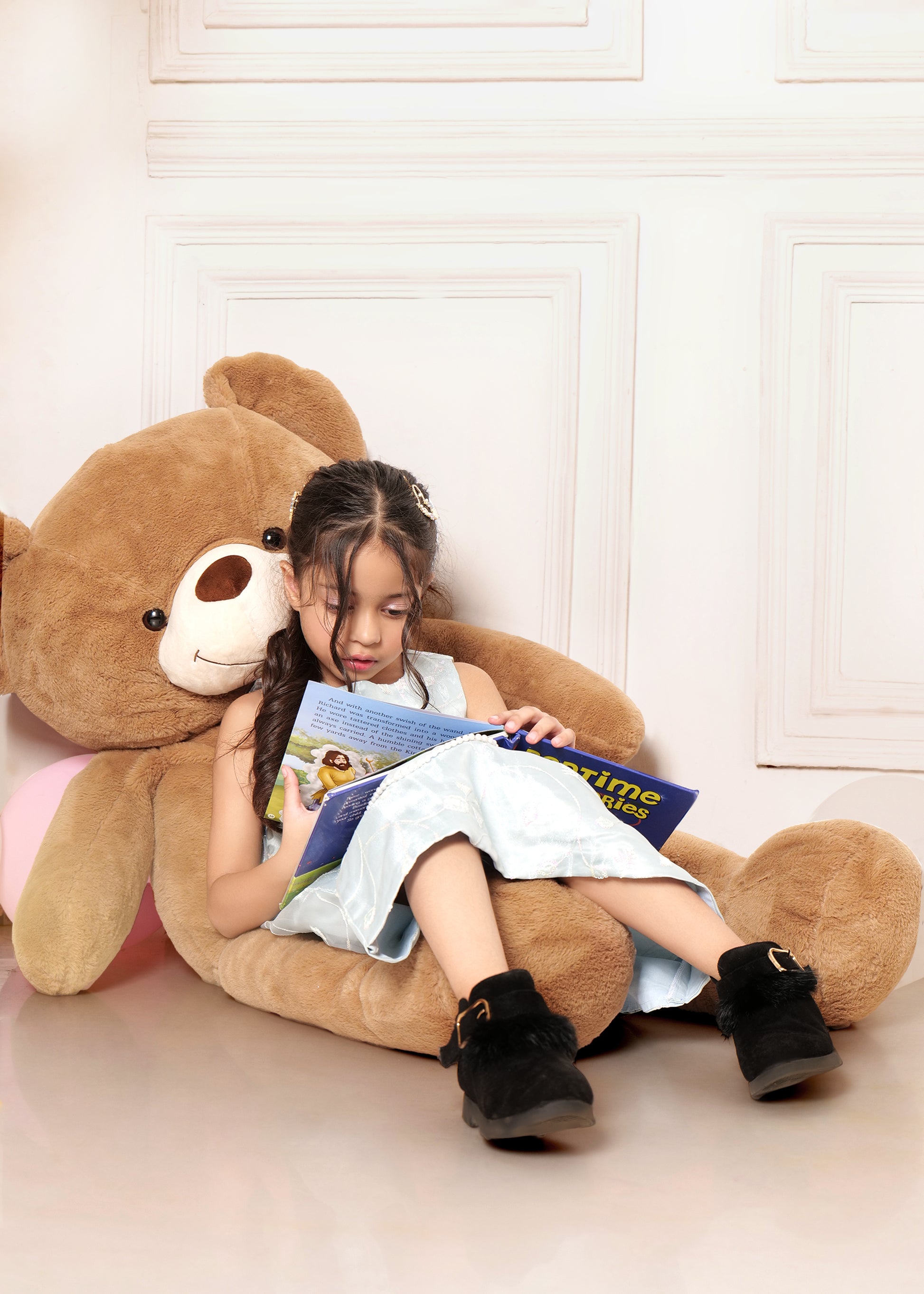 Girl relaxing on a large teddy bear while reading a book, dressed in a light blue embroidered midi dress and black boots, capturing a cozy and playful indoor moment