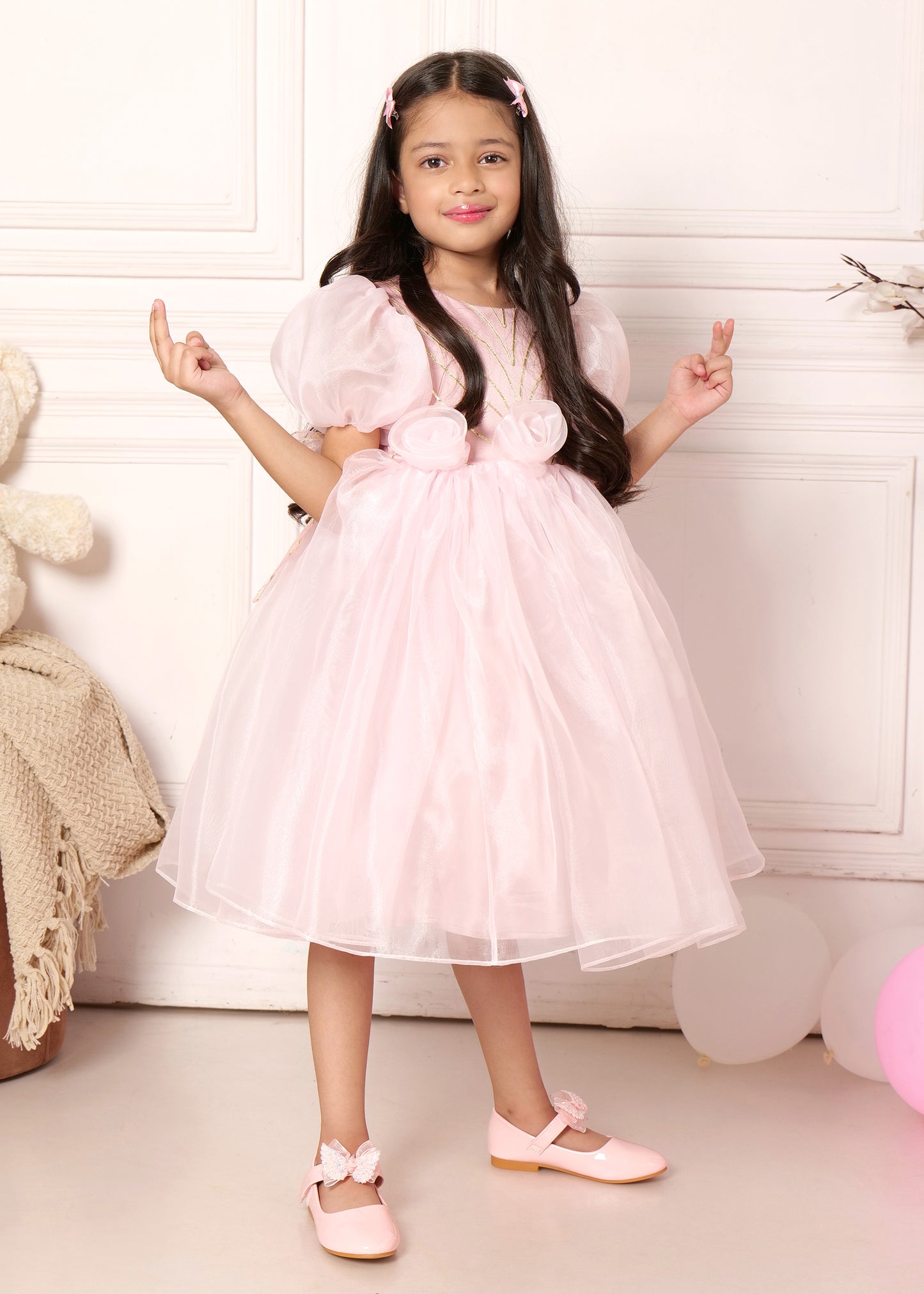 "Young girl in a pastel pink puff sleeve dress with floral designs, striking a pose with delicate curls and matching pink shoes."