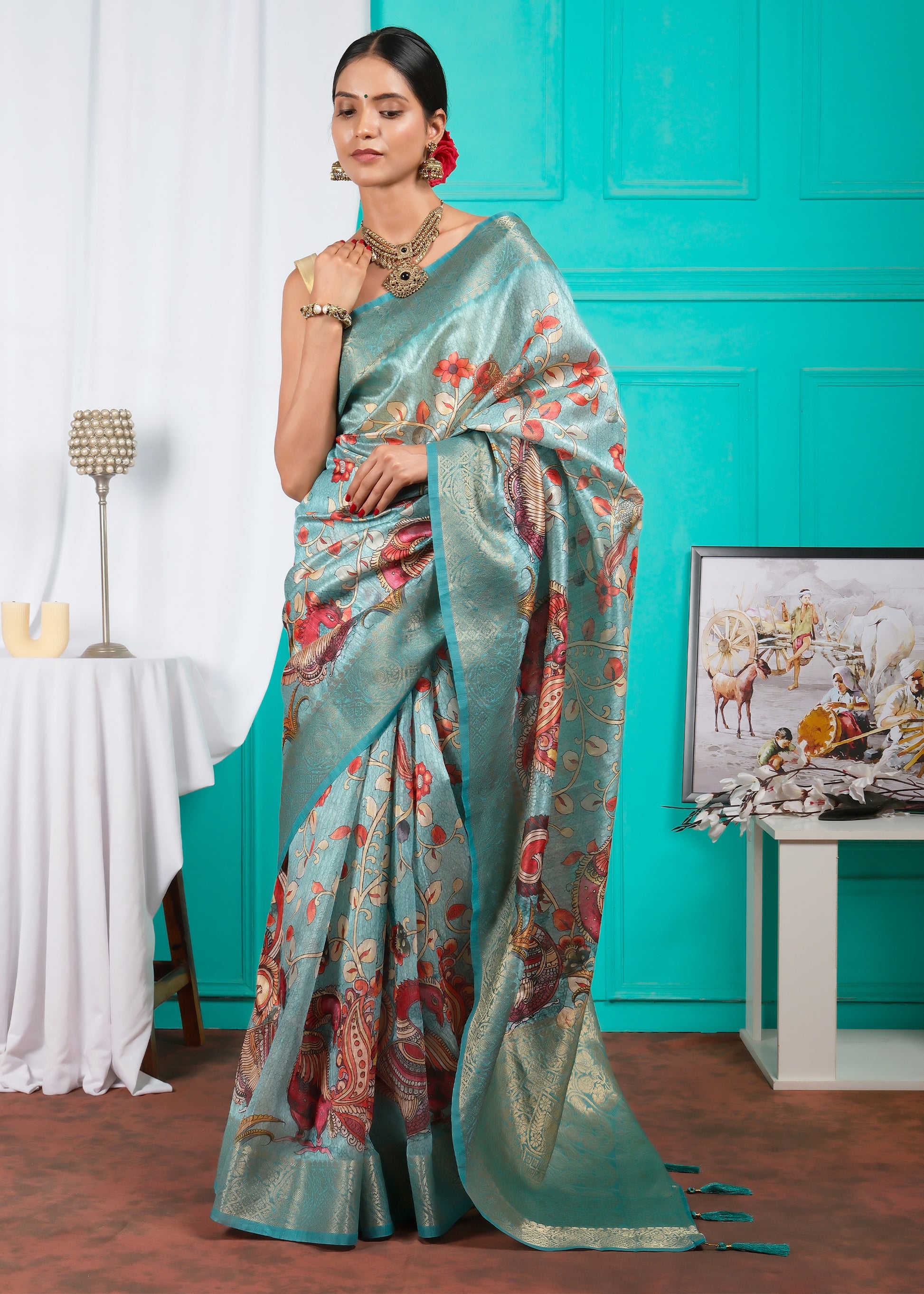 Woman in a teal silk saree featuring peacock and floral designs, posed against a modern backdrop for a festive and elegant look