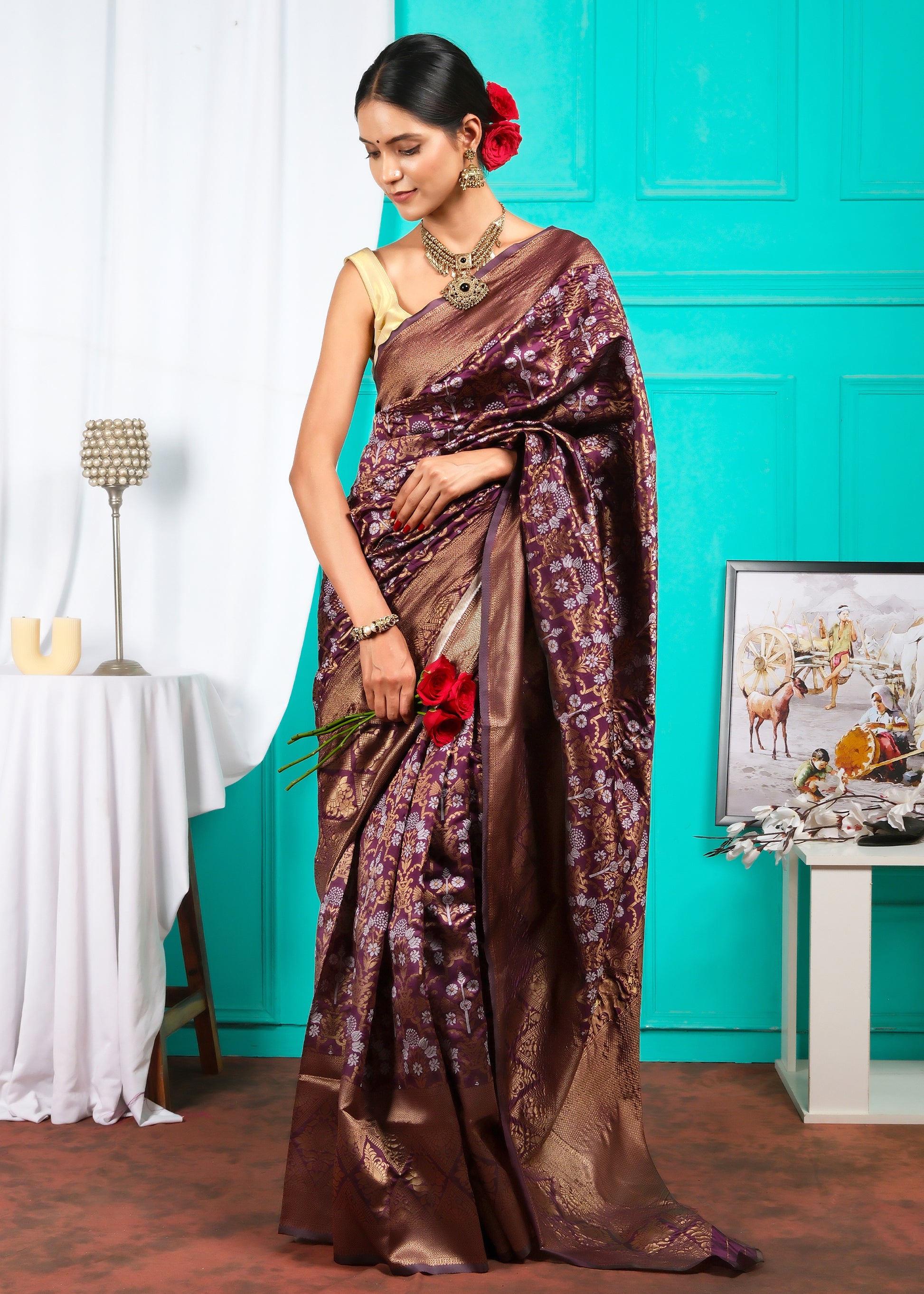  A poised woman sitting while wearing a luxurious maroon and gold saree, accessorized with traditional Indian jewelry, against a vibrant indoor backdrop.