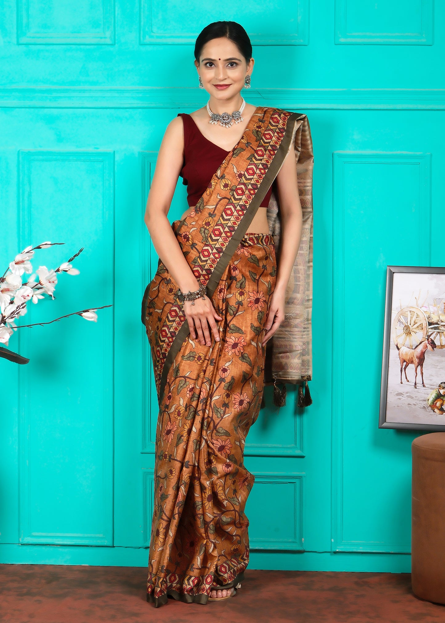 A woman in a traditional Indian saree, posed against a turquoise wall. The saree is adorned with floral patterns in earthy orange tones and features a bold geometric border. She pairs it with a maroon blouse and silver ethnic jewelry, creating a striking look