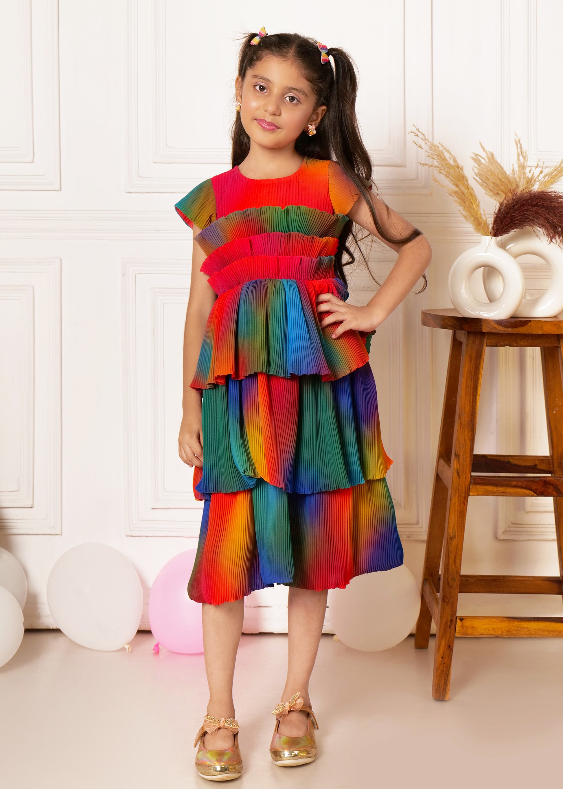 "Girl wearing vibrant rainbow pleated dress, posing confidently with hand on hip, kids occasion wear."