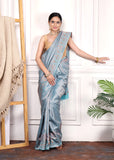 Model posing in a light blue Banarasi silk saree with floral brocade, standing against a neutral background with gold jewelry