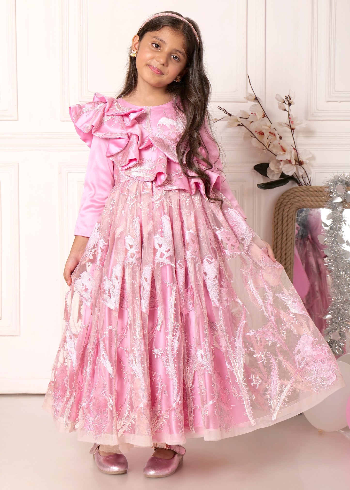 Full-length view of a young girl in a pink gown with long sleeves and ruffled details, set against a decorative background.