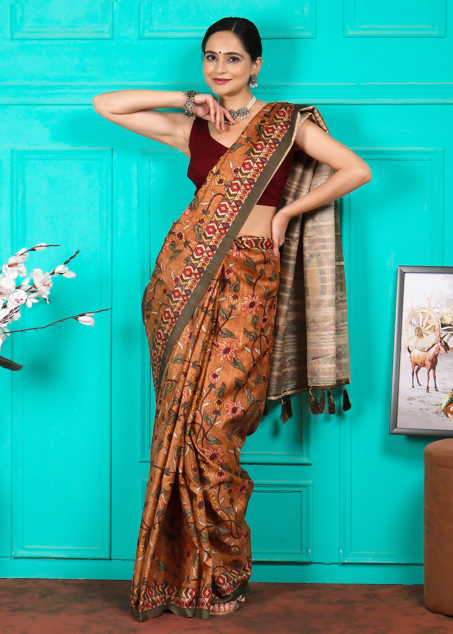 The same woman striking a confident pose in a floral handloom saree, set against a bright turquoise backdrop. The saree's intricate design is complemented by her maroon blouse and silver accessories, giving her a poised and elegant appearance.