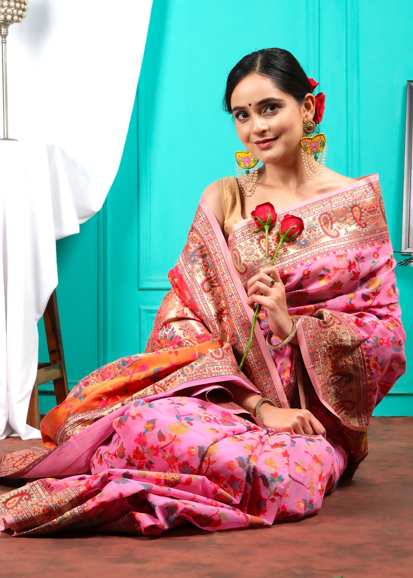 Woman holding roses, sitting elegantly in a pink floral saree with golden embroidery, in a soft traditional pose