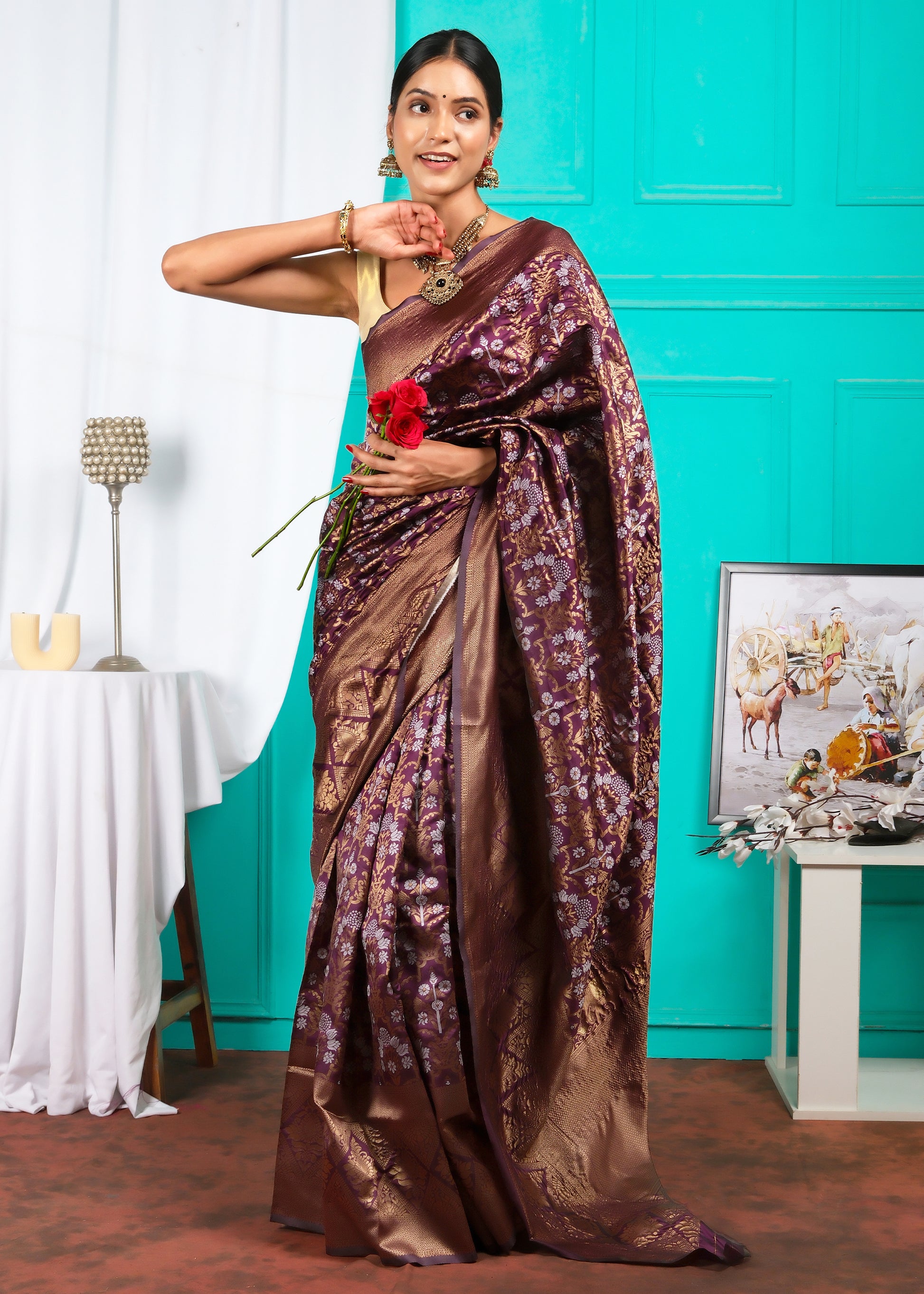 A graceful woman posing in a maroon silk saree with elaborate gold motifs, standing against a colorful backdrop, holding a bouquet of red roses