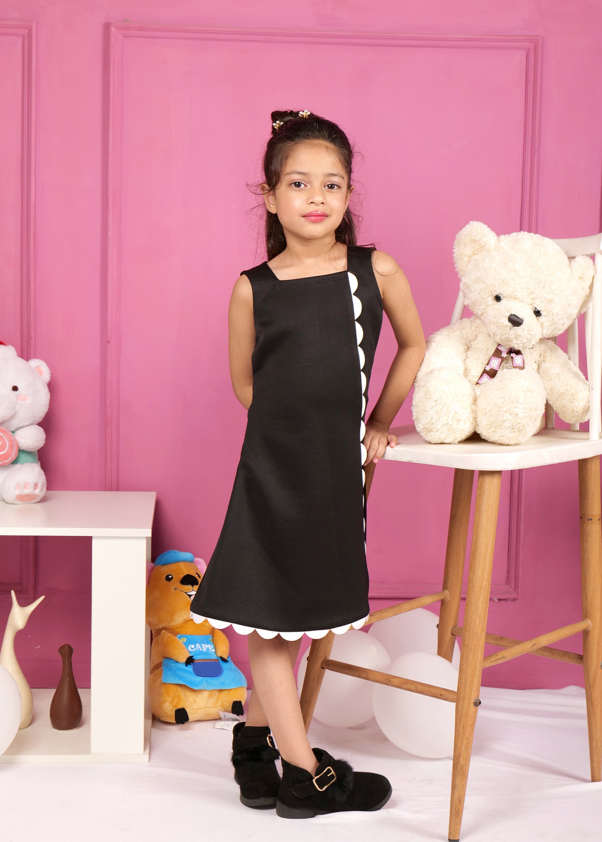 Girl standing in a black dress with white scalloped trim, posing next to a white teddy bear on a table