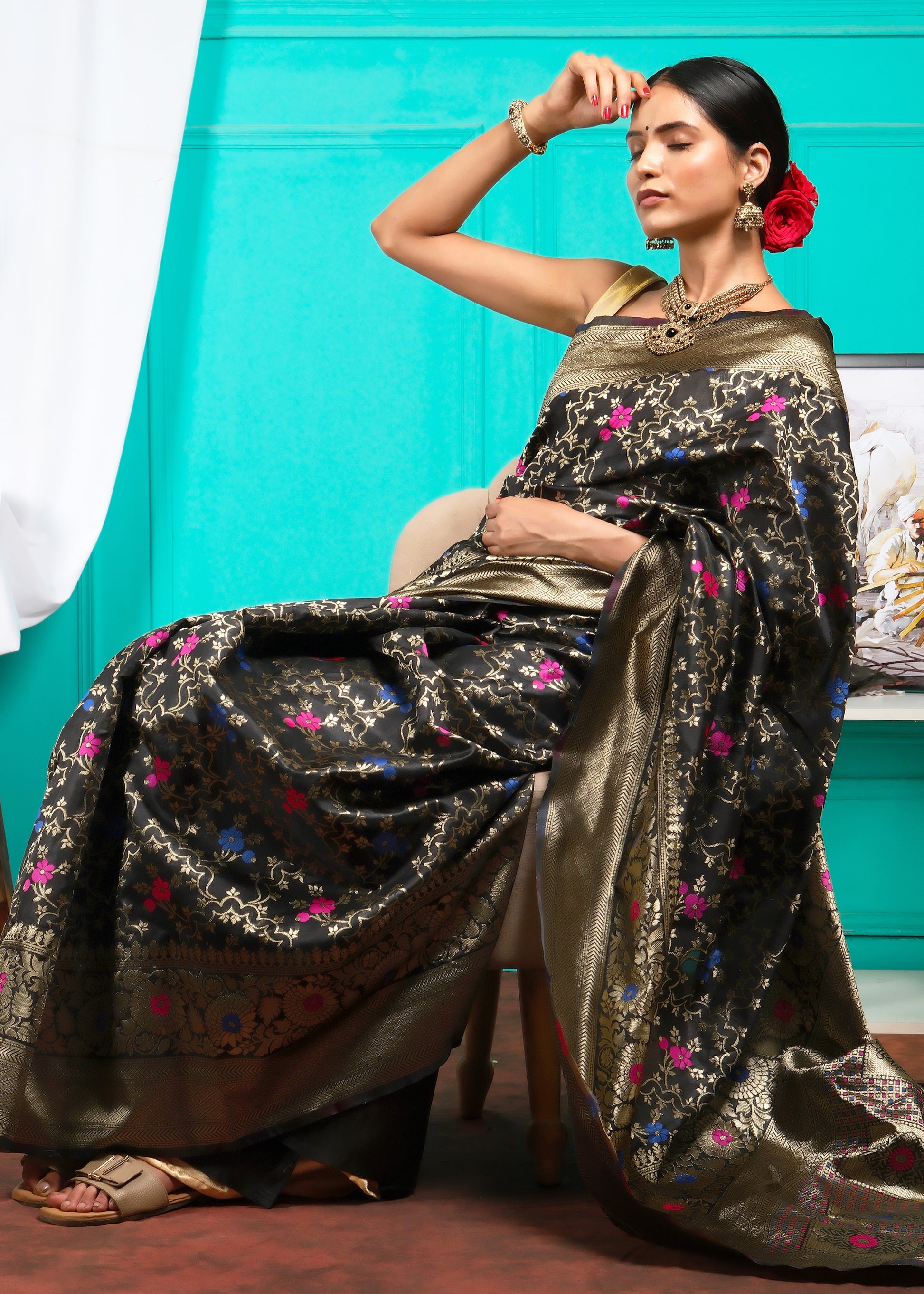 Model posing with a rose while seated in a black Banarasi saree with gold zari work and floral patterns