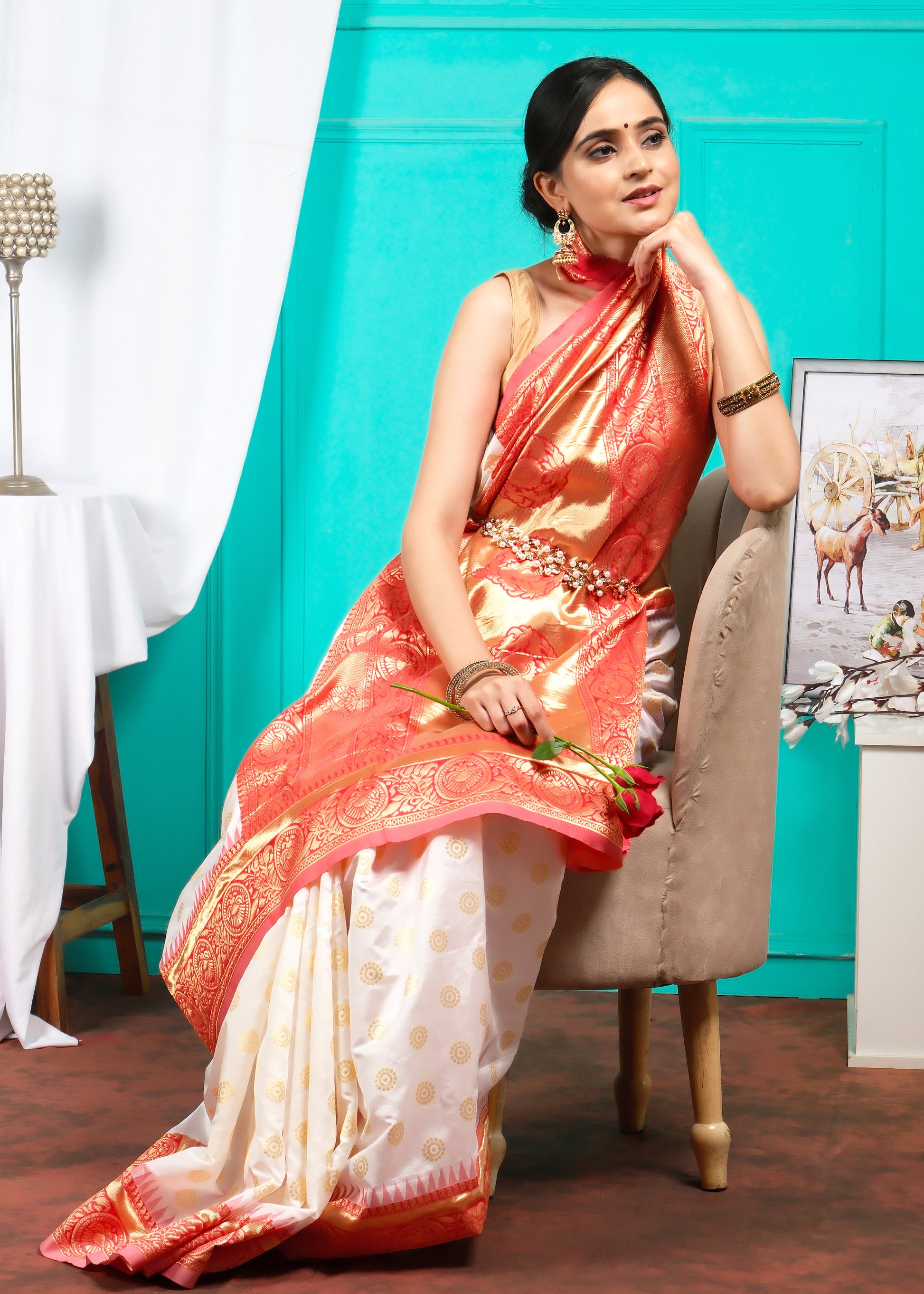 Model gracefully sitting in a traditional white and orange saree with golden designs, against a vibrant background.