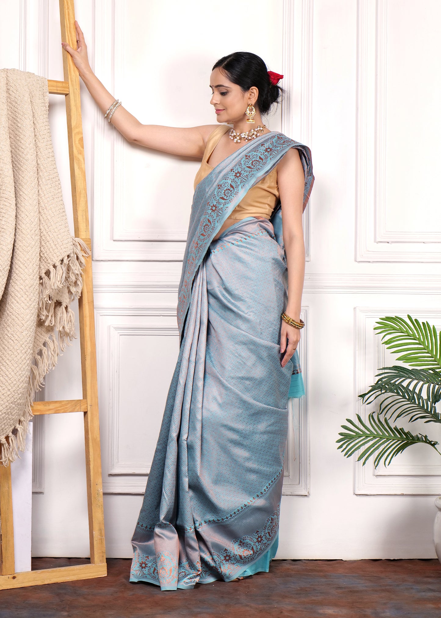 Model showcasing a light blue Banarasi silk saree with intricate border and paired with a sleeveless blouse, leaning against a wooden prop