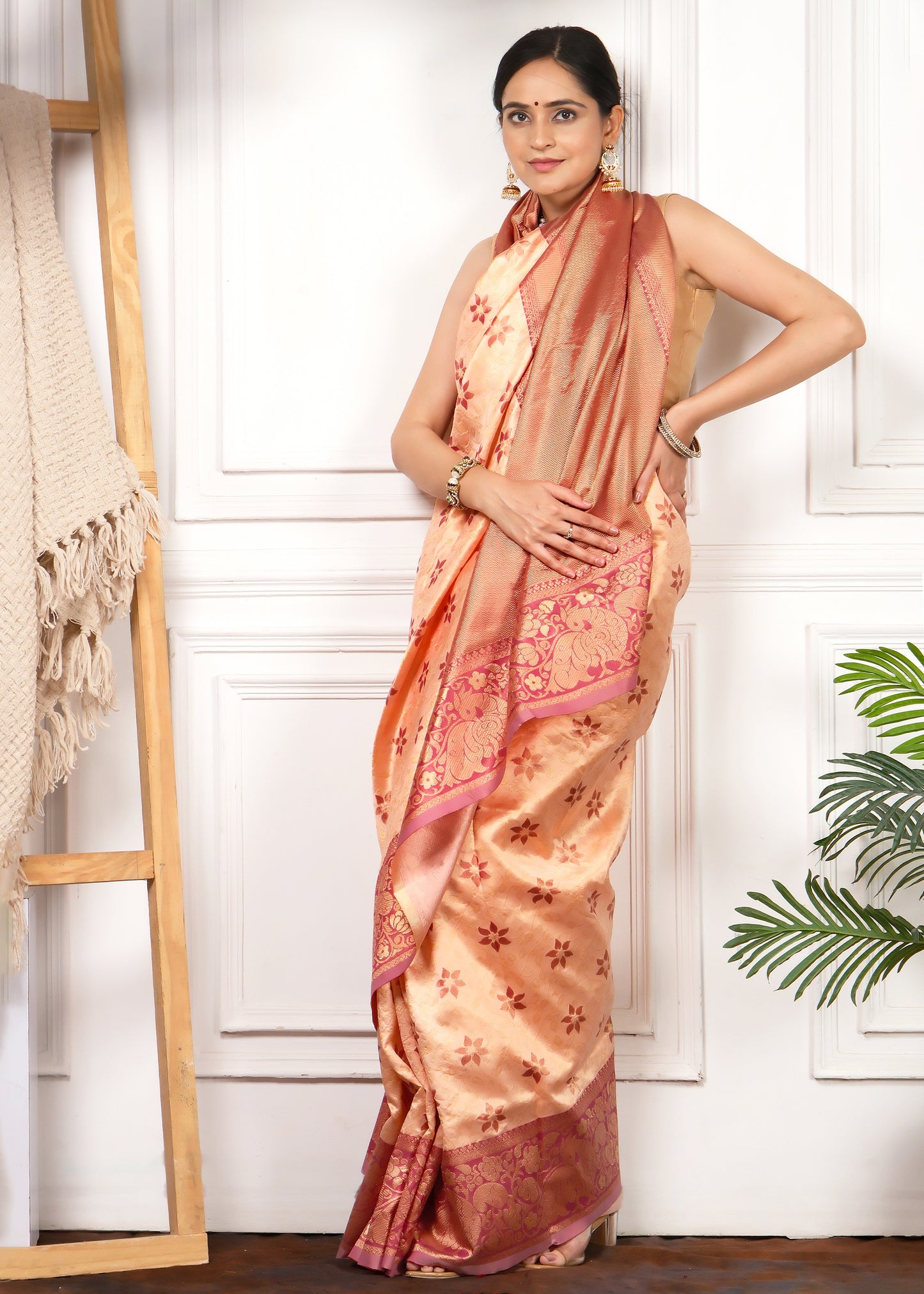 Woman gracefully posing in a peach silk saree with rich gold detailing and a luxurious pink border.