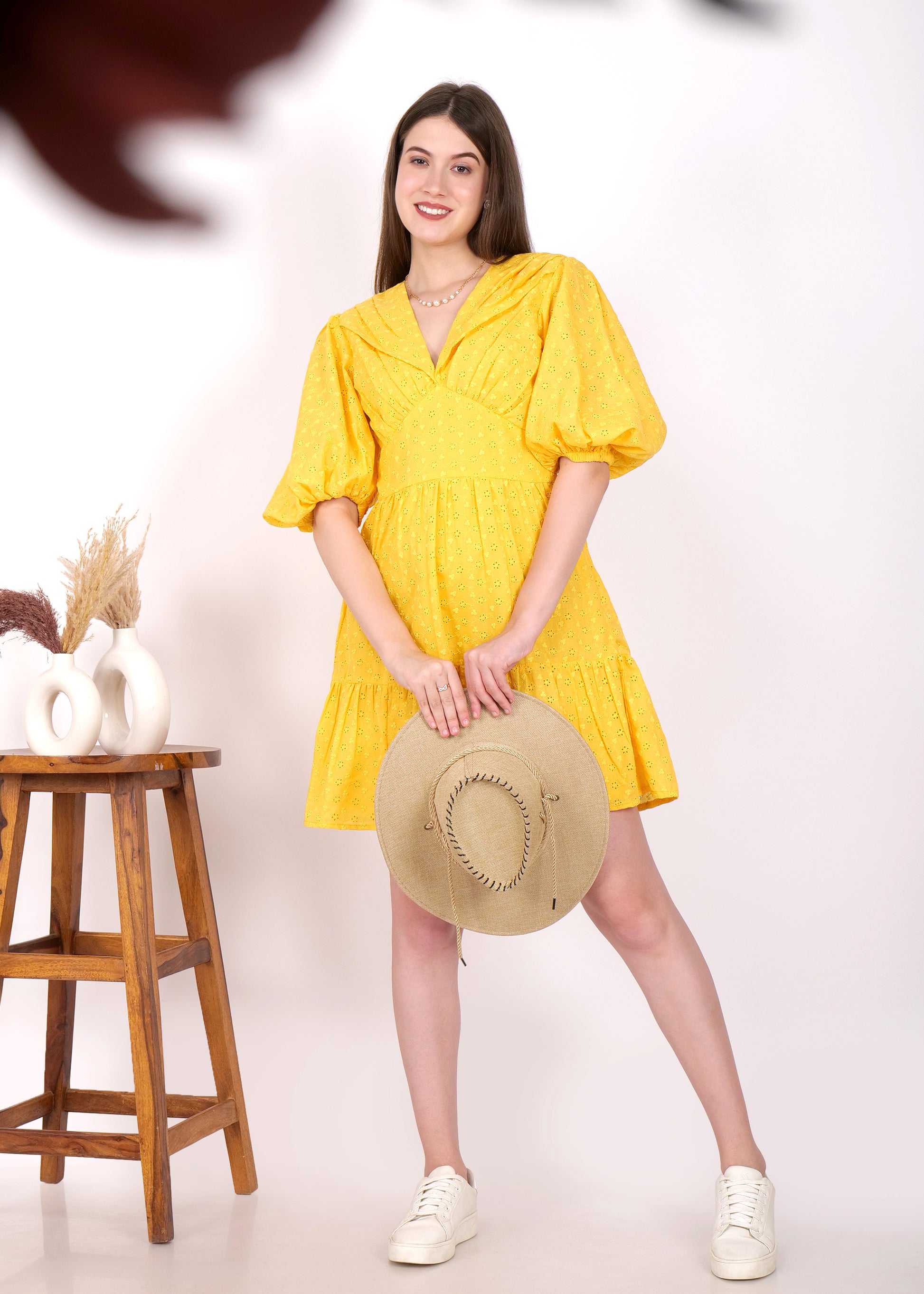 Woman holding a hat while wearing a yellow puff sleeve dress and white sneakers, presenting a laid-back summer outfit in a studio setup