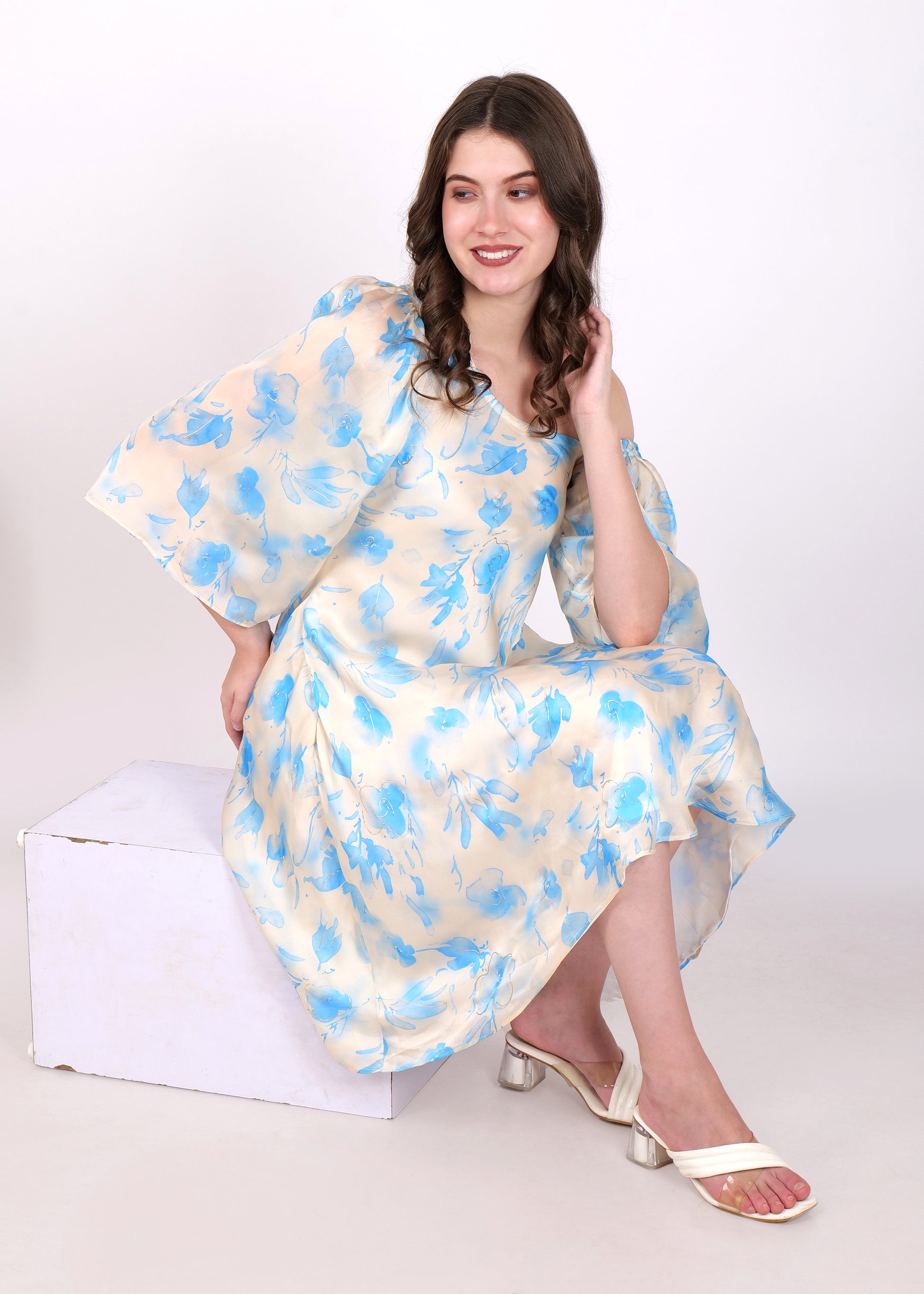 Woman sitting and smiling while modeling a cream-colored floral dress with blue flowers