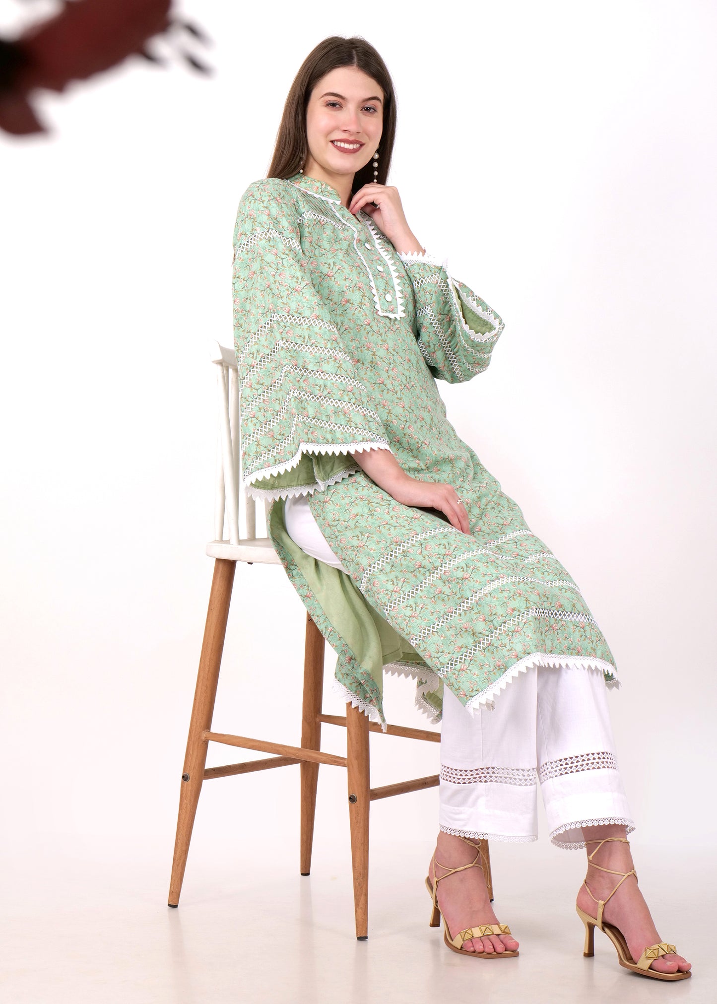 Woman sitting on a chair, wearing a green floral print kurta with lace accents, paired with white pants and strappy heels