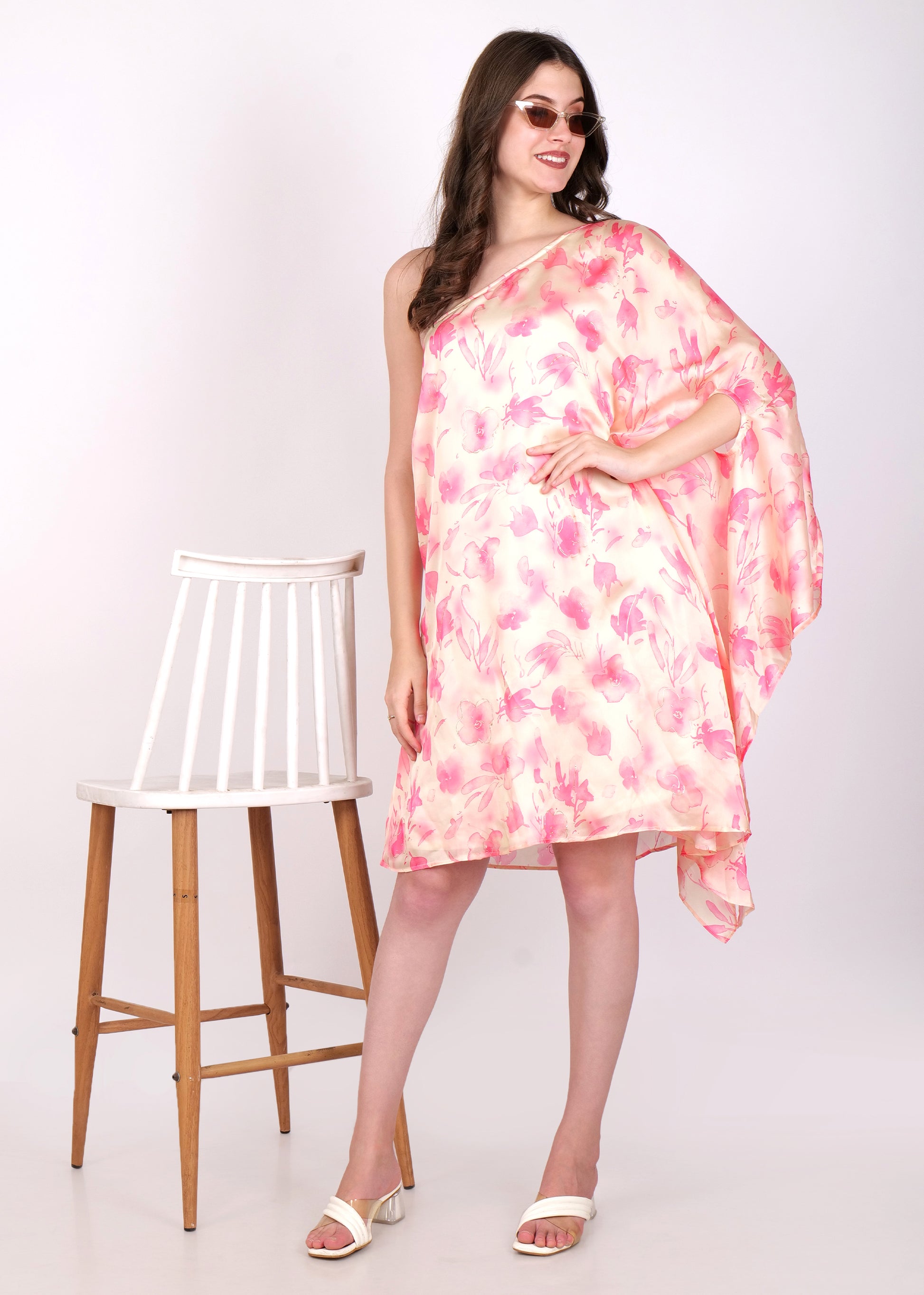 Model wearing a pink and peach watercolor floral dress paired with sunglasses, showcasing a summer-ready look with a chair as a prop