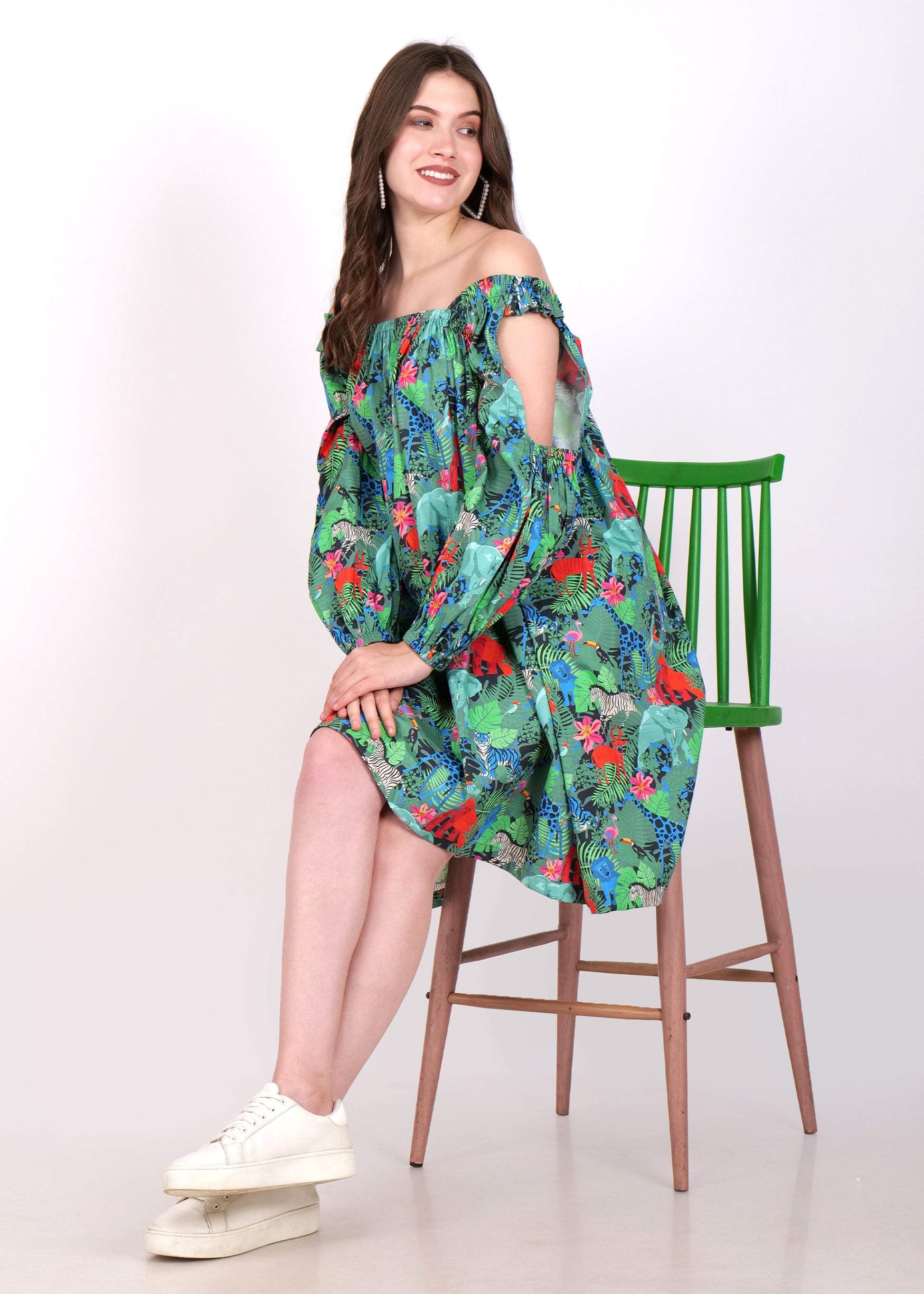 Woman sitting on a green chair, modeling a jungle-printed, off-shoulder dress with stylish cutout sleeves and casual white sneakers