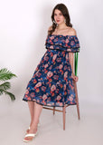 Woman seated in a navy blue off-shoulder floral pleated midi dress, featuring ruffled layers and lace trim, styled with white sandals