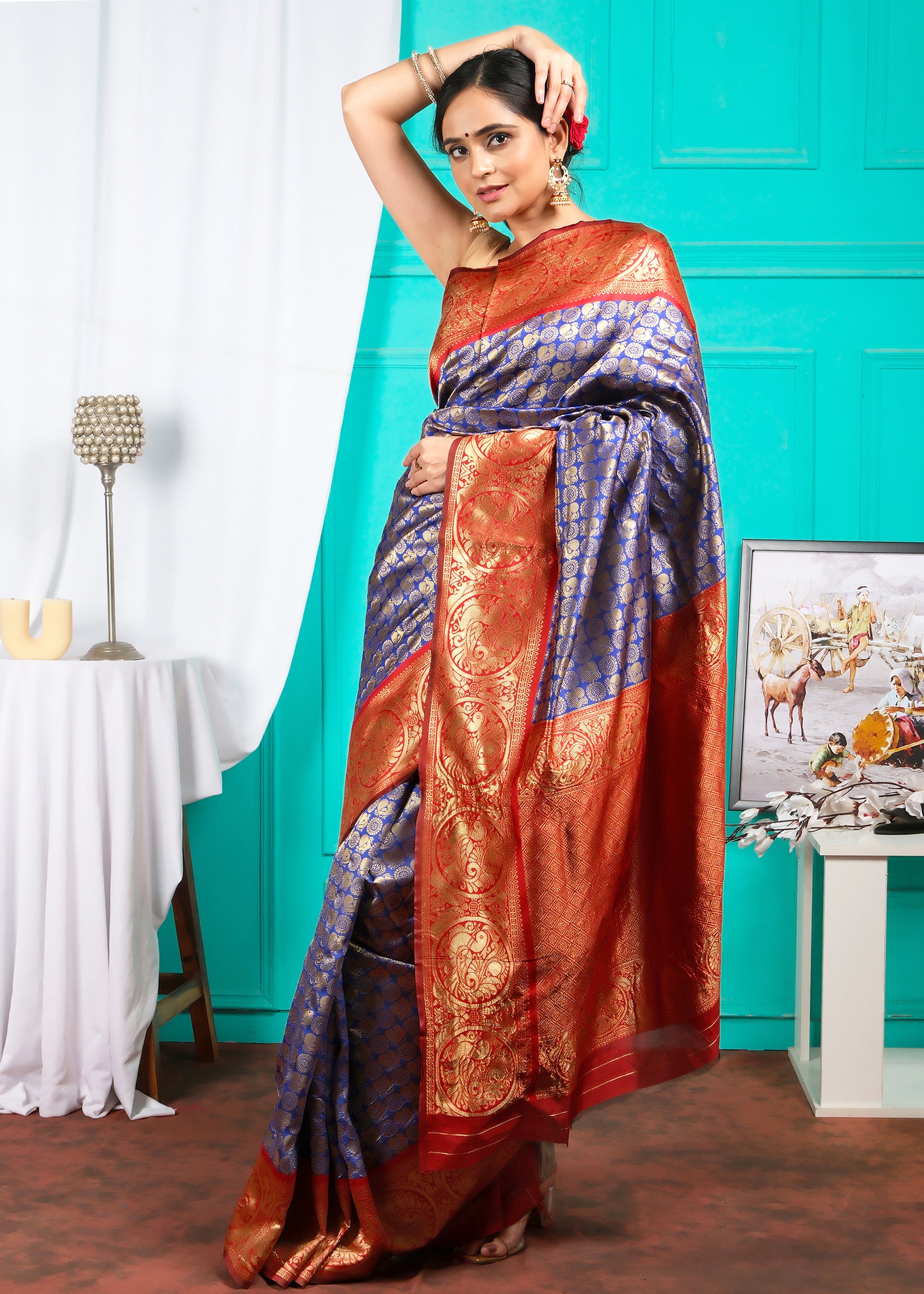 A woman gracefully posing in a royal blue and red Banarasi saree, featuring golden zari work and a rich textured pallu