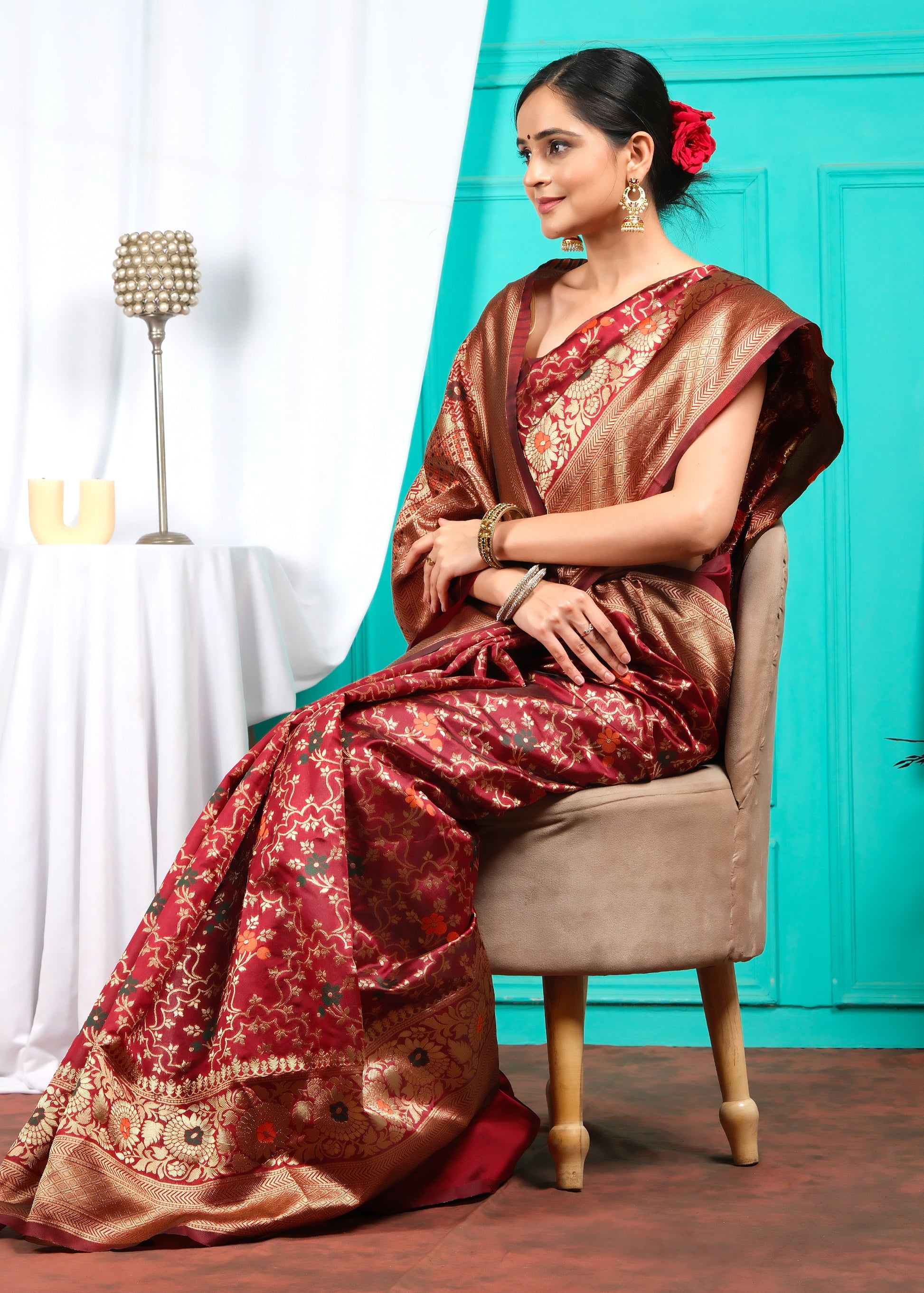 Graceful woman in a red Banarasi saree, showcasing the rich floral designs and golden zari border while seated on a chair