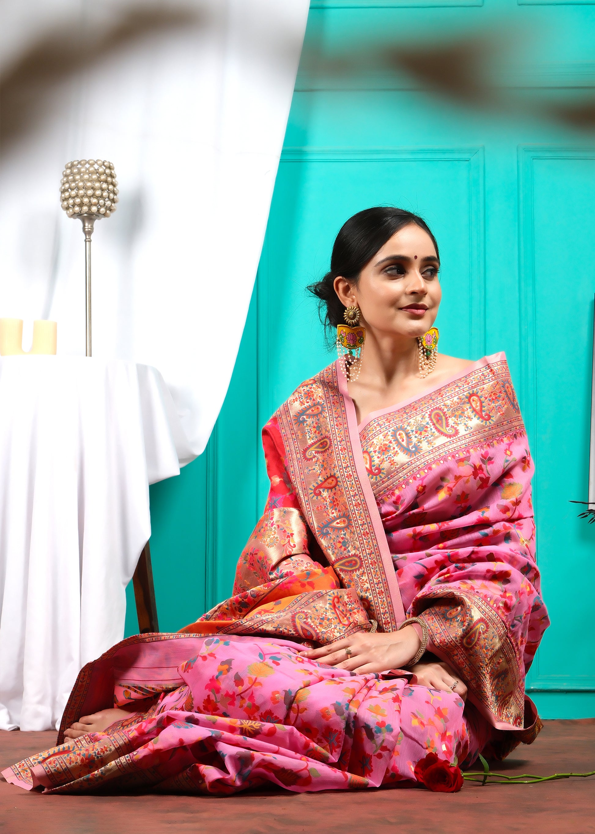 Woman posing serenely in a pink floral saree with intricate detailing and bold paisley patterns, sitting on the floor.