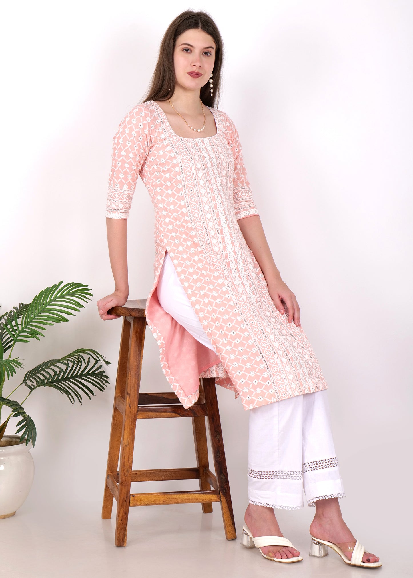 Model posing next to a stool, wearing a pink embroidered cotton kurta with white lace trim and white trousers