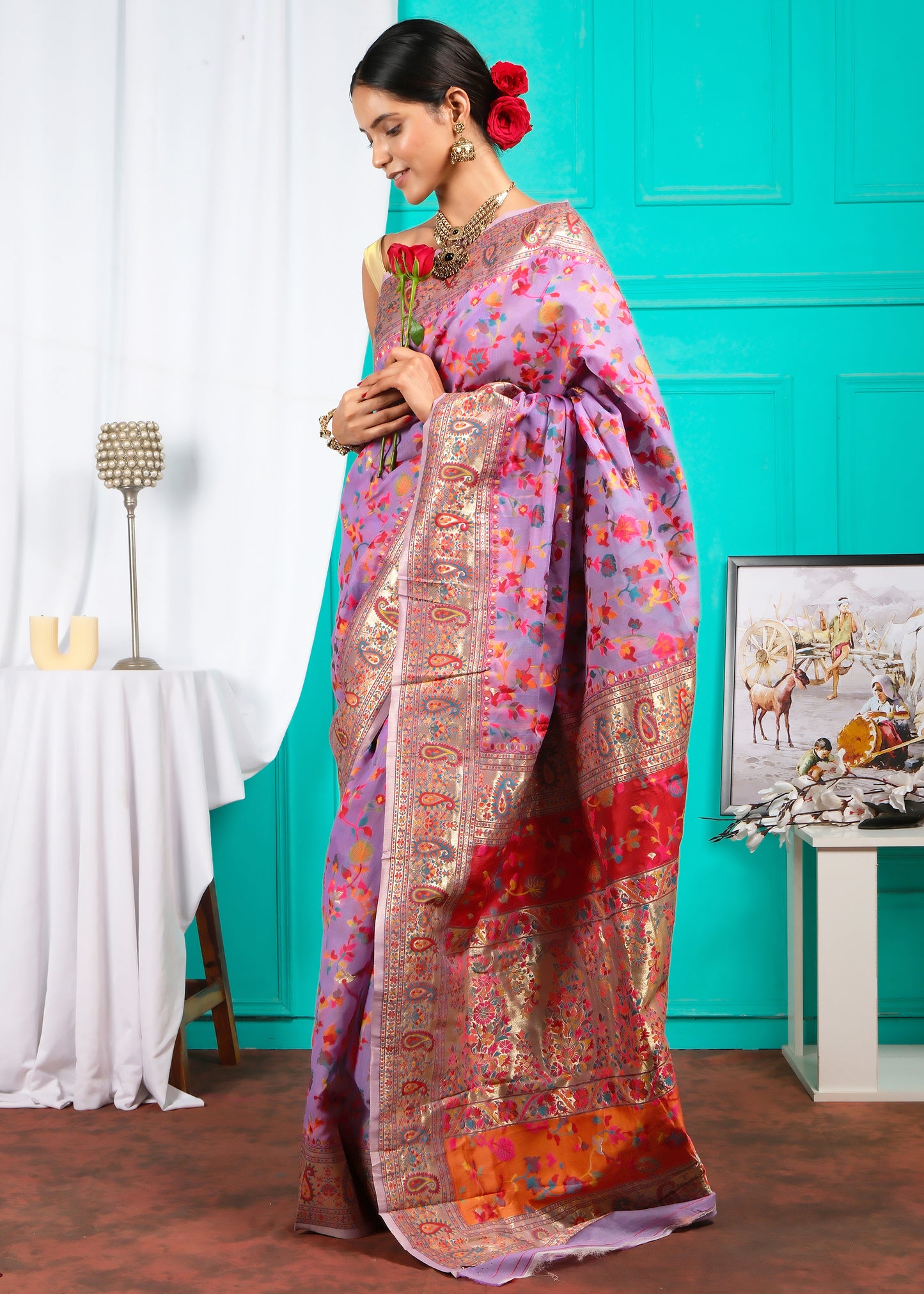 Woman in a lavender floral print saree, standing with a graceful posture, featuring a contrasting red and gold intricate pallu and ornate accessories