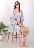 Model sitting with sunglasses and a hat, styled in a watercolor floral off-shoulder dress