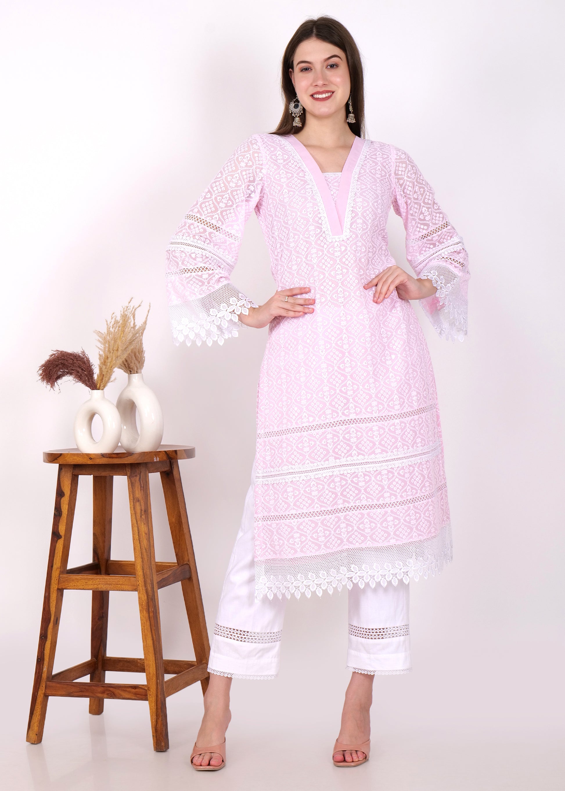 Woman wearing a pastel pink embroidered kurta with lace sleeves, styled with white pants, standing beside a wooden stool with decorative vases.