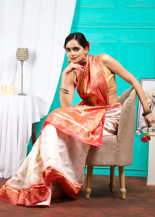 Model posing in a seated position, draped in a luxurious white and orange silk saree, exuding elegance and charm