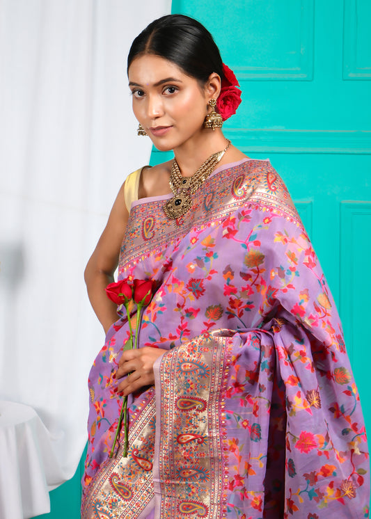 Woman in lavender saree adorned with vibrant floral motifs and a red rose, posing elegantly against a teal background, showcasing intricate jewelry and cultural elegance