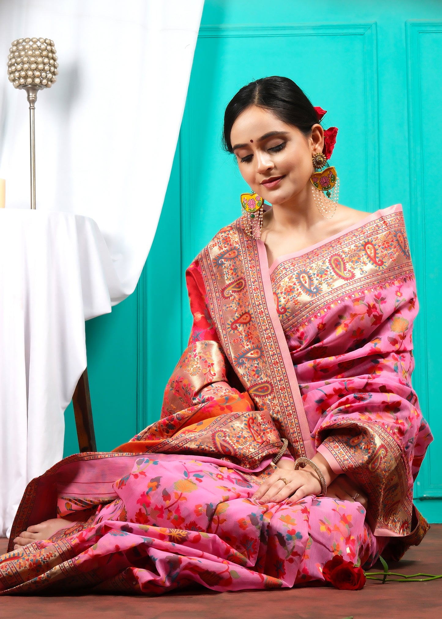 Woman gazing softly, draped in a pink saree with floral motifs, seated in a traditional setting