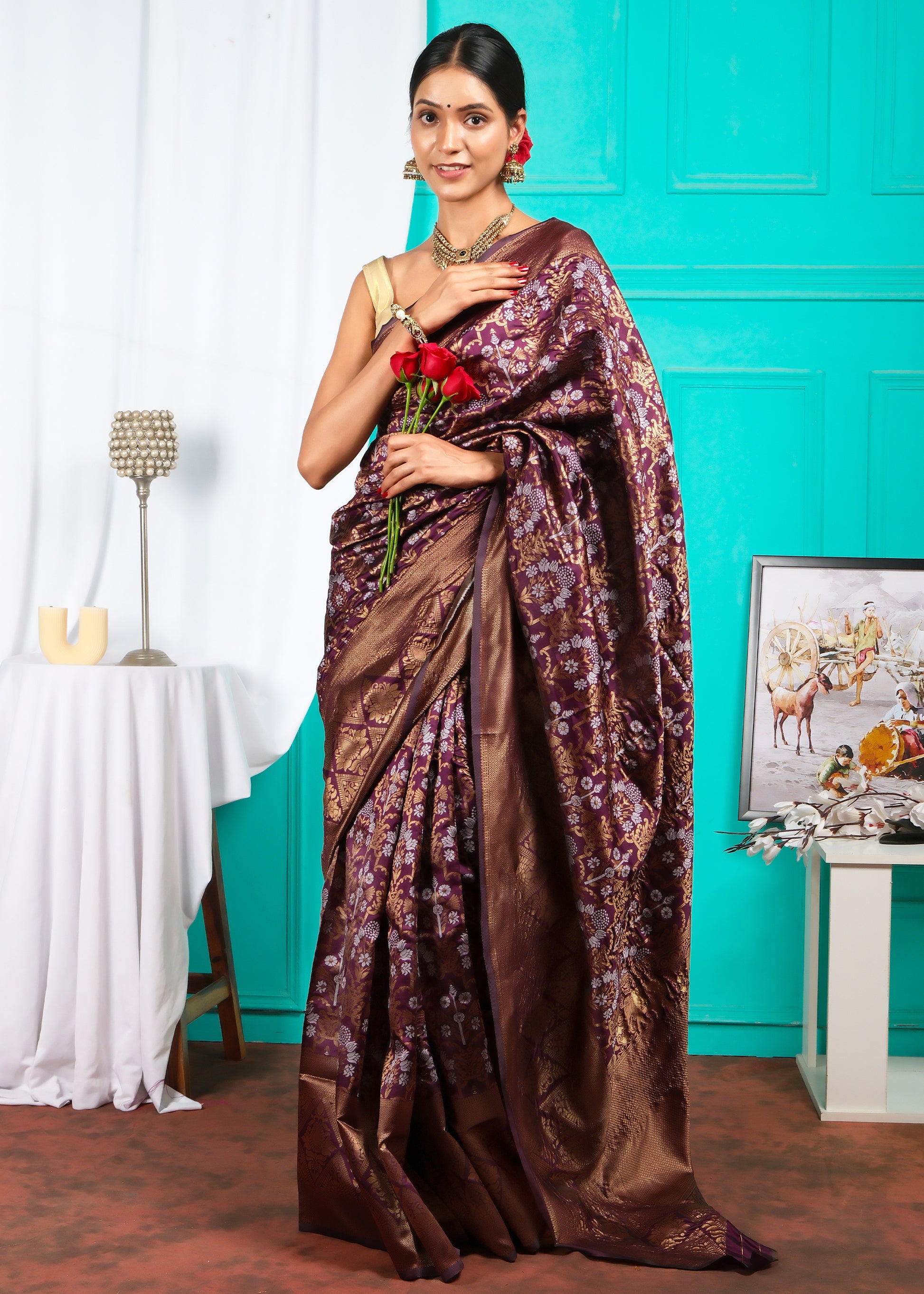 A stylish woman draped in a maroon silk saree, adorned with beautiful gold floral patterns, holding roses and posing in front of a colorful background