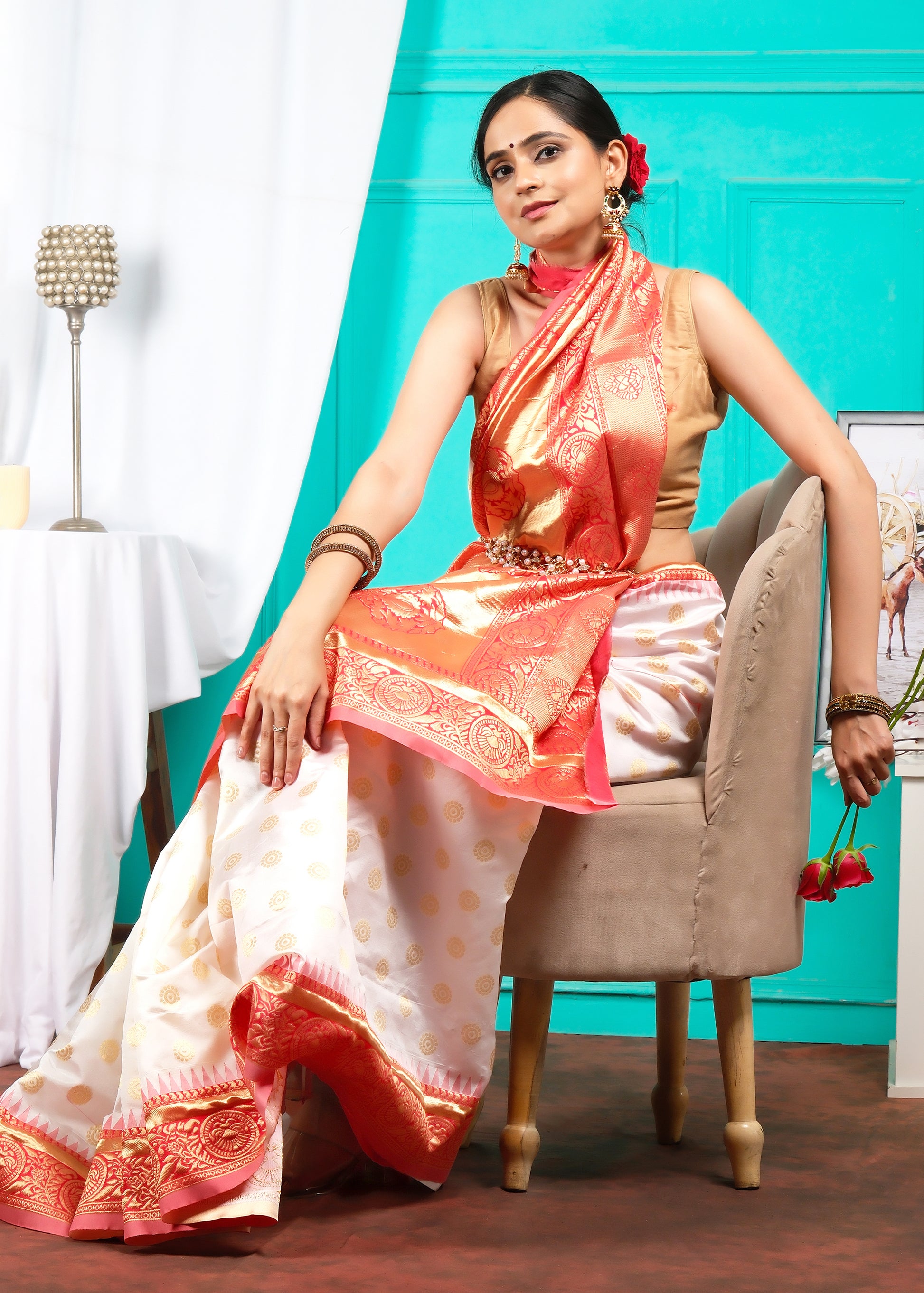 Fashionable portrayal of a model wearing a white and orange silk saree with golden motifs, seated stylishly with flowers