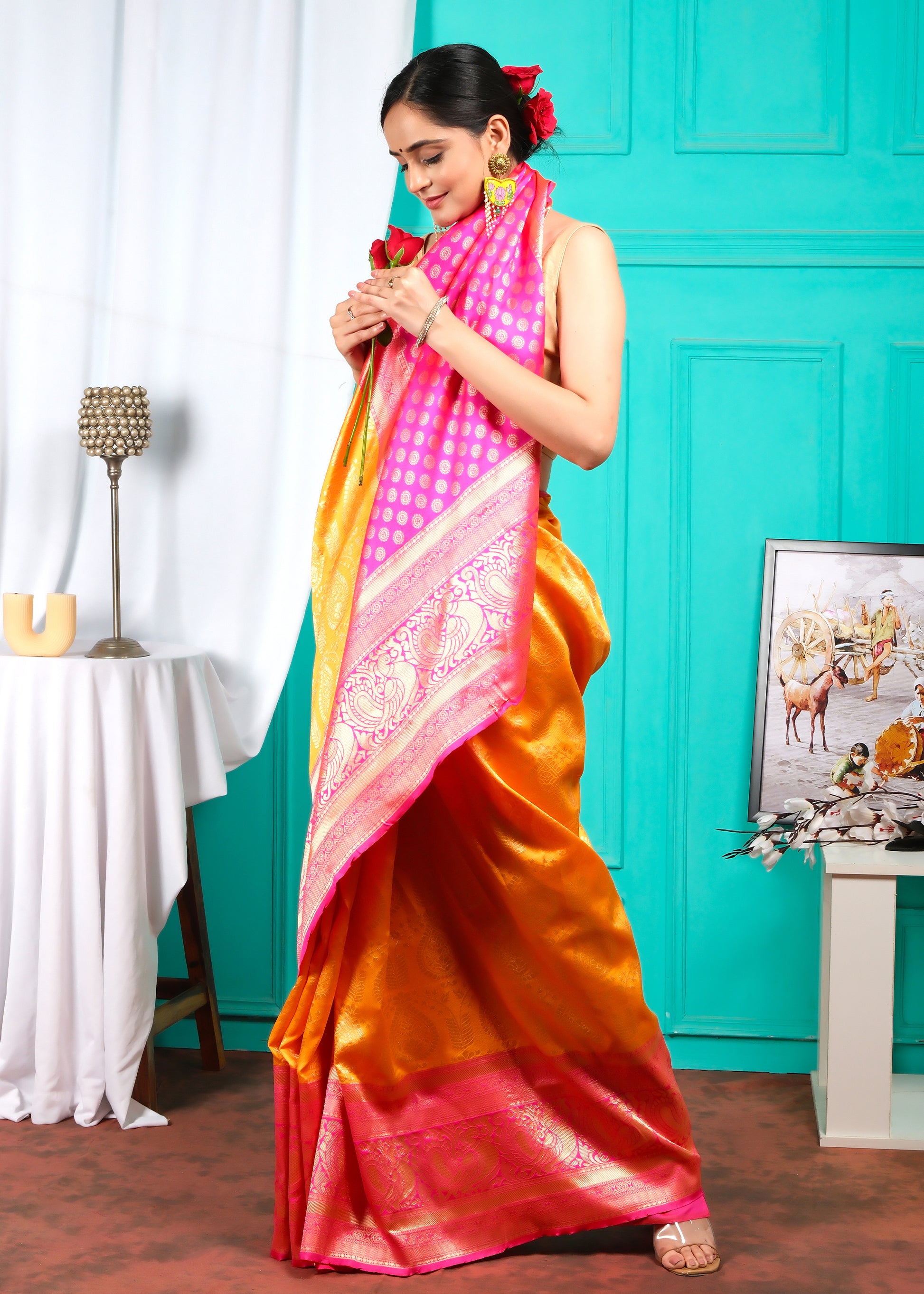 Woman smiling in an orange and pink silk saree, holding a rose, with intricate golden motifs and a regal pink border, perfect for festive occasions